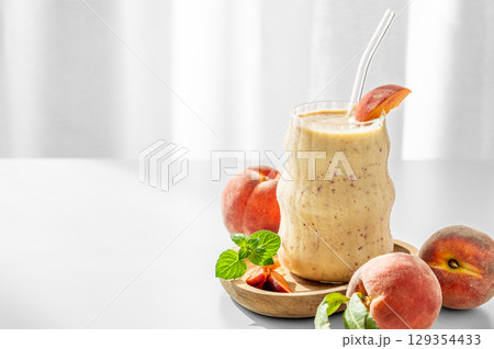 Smoothie with peach and banana in a glass on a wooden board on a white table near window Smoothie with peach and banana in a glass on a wooden board on a white table near window 129354433