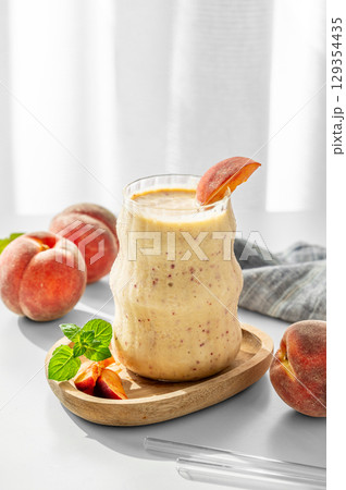 Smoothie with peach and banana in a glass on a wooden board on a white table near window Smoothie with peach and banana in a glass on a wooden board on a white table near window 129354435