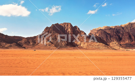 Flat red desert with large mountains in distance, blue sky above, some off road vehicles in background, typical scenery at Wadi Rum, Jordan 129355477
