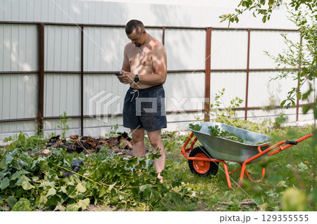 Shirtless Man Gardening Outdoors with Wheelbarrow, Brown Shorts, Green Foliage, Metal Fence, Sunny Day, Back Yard, Natural Light Photography 129355555