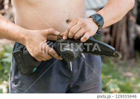Man Adjusting Chainsaw Battery Outdoors, Focus on Tool Handling, Greenery Background, Black and Grey Tones, Summer Maintenance, Waist-Up Shot, Natural Lighting 129355646