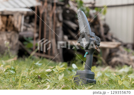 Electric Cordless Handheld Chainsaw Cutting Small Branches Outdoors, Green Grass, Rustic Wood Pile, Garden Tool, Power Equipment, Sharp Blade Electric Cordless Handheld Chainsaw Cutting Small Branches Outdoors, Green Grass, Rustic Wood Pile, Garden Tool, Power Equipment, Sharp Blade 129355670