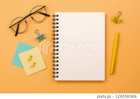 Notebook and stationery on orange background. Blank spiral notebook with pen, glasses, push pins and binder clips 129356369