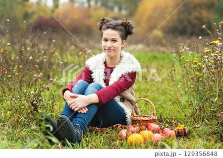 Thanksgiving Vibes. Young girl sitting on blanket in autumn meadow holding two small pumpkins, surrounded by wildflowers. warmth and whimsy of autumn harvest, childhood, and outdoor seasonal fun 129356848