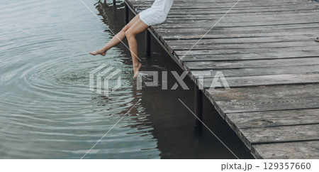 unrecognizable womans legs in a white dress, sitting on the edge of wooden pier, dipping her feet into calm water with a light splash, relaxation, travel 129357660