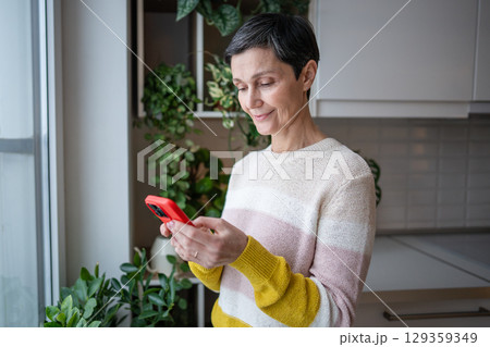 Content happy cheerful mature woman smiles happily, surfing internet chatting smartphone in kitchen 129359349