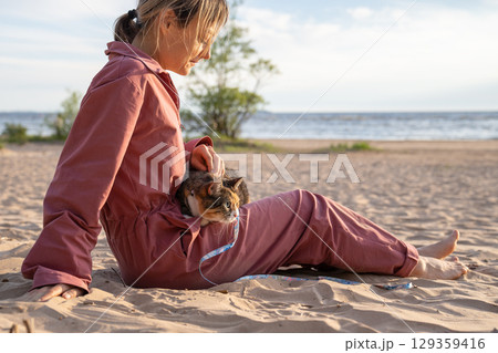 Lively curious cat watching sandy beach held by owner, woman lies down petting feline on shore Lively curious cat watching sandy beach held by owner, woman lies down petting feline on shore 129359416