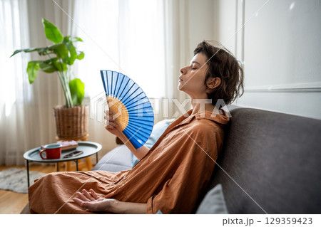 Exhausted sweaty woman fanning herself sitting on sofa suffering from summer heat without AC 129359423