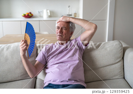 Older man struggling with fan heat exhaustion. Pensioner try to cope in sweltering home in summer Older man struggling with fan heat exhaustion. Pensioner try to cope in sweltering home in summer 129359425