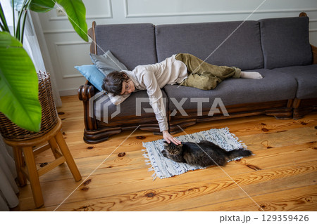 Tired woman resting at home petting cat friend lying beside couch on soft rug at home, slow life 129359426