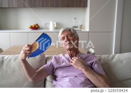 Overheated senior man sits on home couch waving paper fan to cool down, suffers from stuffiness 129359434