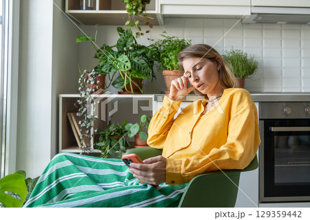 Thoughtful serious woman scroll news and social feeds on phone surrounded by indoor plants at home Thoughtful serious woman scroll news and social feeds on phone surrounded by indoor plants at home 129359442