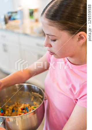 A girl in a pink shirt stirs cookie dough filled with chocolate candies in a stainless steel bowl. She is working at a kitchen counter in a well-lit, modern home. 129359588