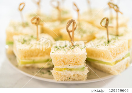 Small tea sandwiches made with soft white bread, layered with cream cheese spread, thinly sliced cucumber, and topped with fresh dill. Each sandwich is secured with a bamboo cocktail pick and served Small tea sandwiches made with soft white bread, layered with cream cheese spread, thinly sliced cucumber, and topped with fresh dill. Each sandwich is secured with a bamboo cocktail pick and served 129359603