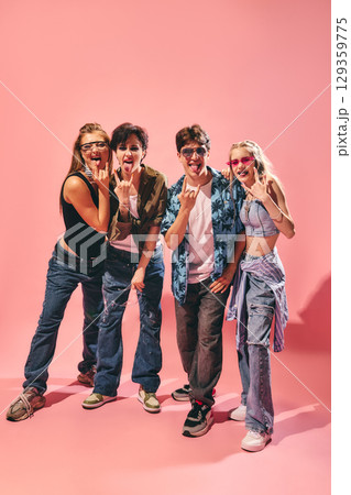Group of happy young friends showing rock signs and sticking tongues out on pink background in casual outfits Group of happy young friends showing rock signs and sticking tongues out on pink background in casual outfits 129359775