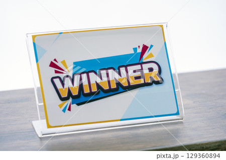 Colorful sign with word WINNER displayed on clear stand, symbolizing success and achievement. vibrant design includes geometric shapes and dynamic lines, creating energetic feel Colorful sign with word WINNER displayed on clear stand, symbolizing success and achievement. vibrant design includes geometric shapes and dynamic lines, creating energetic feel 129360894