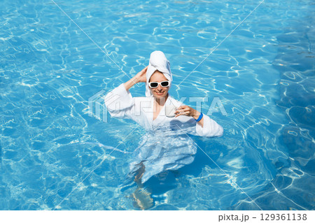 A girl in a white coat swims in a blue pool. Wearing sunglasses and a towel. Epilation of legs, varicose veins. Rest, relaxation on the beach, at the hotel. A woman in freedom. Vacation. High quality 129361138