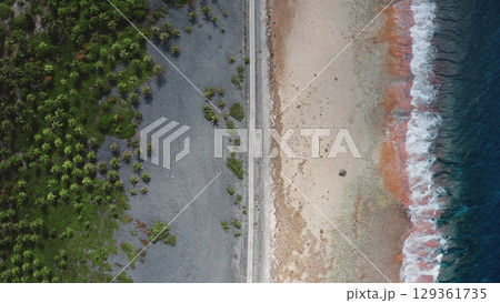 French Polynesia, Rangiroa island: Aerial view capturing a thin road separating lush greenpalm trees vegetation from sandy beach and crashing ocean waves. Drone flight top down 129361735
