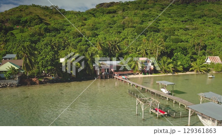 Tranquil lagoon waters lapping wooden pier lead to cozy beachfront houses nestled among lush tropical vegetation and a majestic verdant hillside. Tahiti tropical island, French Polynesia. Drone flight 129361863