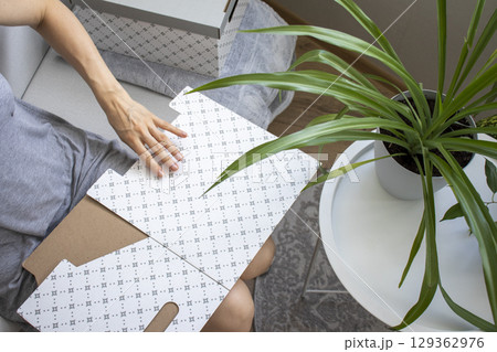 Woman sitting in the couch in cozy room, assembling carton box. Indoors Woman sitting in the couch in cozy room, assembling carton box. Indoors 129362976