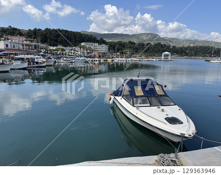 Coastal village in Greece showcases tranquil harbor with boats gently bobbing on calm waters against backdrop of lush hills and vibrant sky, inviting visitors to enjoy seaside charm 129363946