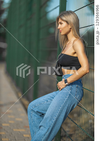 young woman stands near a green fence, sporting a black crop top and blue high waisted jeans She appears relaxed and confident in the bright sunlight, enjoying the outdoor atmosphere young woman stands near a green fence, sporting a black crop top and blue high waisted jeans She appears relaxed and confident in the bright sunlight, enjoying the outdoor atmosphere 129366966