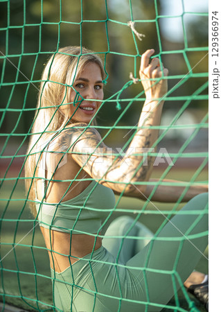young woman with long hair and tattoos enjoys her time at a sports field, sitting behind a green netting and smiling at the camera under bright sunlight 129366974