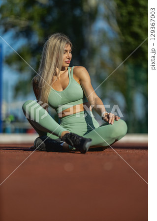 A fitness enthusiast sits cross legged on a track at a sports facility, enjoying the sunny weather while engaging in stretching The scene captures her athletic attire and relaxed posture A fitness enthusiast sits cross legged on a track at a sports facility, enjoying the sunny weather while engaging in stretching The scene captures her athletic attire and relaxed posture 129367003