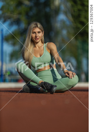 A woman in green activewear is sitting on a running track, exuding confidence and strength She enjoys the sunny weather while showcasing her athleticism and fitness lifestyle 129367004