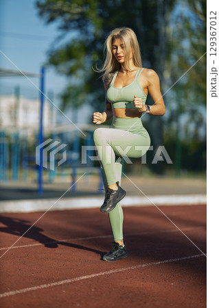 sporty girl is actively jogging in place on running track surrounded green trees She is focused on her fitness routine under clear blue sky creating vibrant atmosphere 129367012