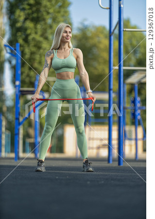 sporty girl stands confidently in a fitness park, stretching a red elastic resistance band Dressed in a mint green outfit, she showcases her athletic physique amidst vibrant green trees 129367018