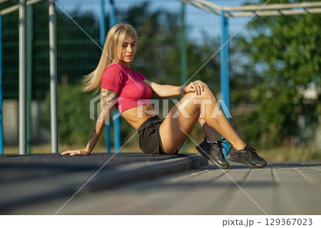 fitness enthusiast sits on a wooden platform at an outdoor gym, enjoying the warm light of late afternoon She wears a fitted top and shorts, showcasing her tattoos and athletic physique 129367023