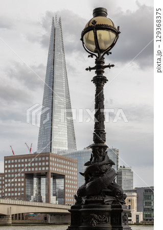 "Dolphin lamp standard" adorned with ornate details in the foreground, a striking, glass-clad skyscraper known as "The Shard" stands tall for its distinctive pointed spire with cloudy sky. 129368375