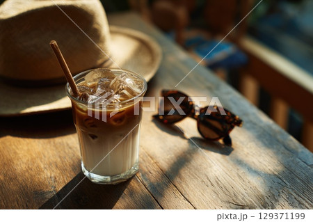 cold coffee drink with ice cubes in an iced latte glass, placed on the wooden table next to sunglasses and a sun hat. 129371199
