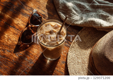 cold coffee drink with ice cubes in an iced latte glass, placed on the wooden table next to sunglasses and a sun hat. cold coffee drink with ice cubes in an iced latte glass, placed on the wooden table next to sunglasses and a sun hat. 129371200