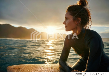A female surfer in her wetsuit, sitting on a surfboard at sea with a sunset in the background 129371258