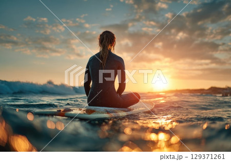 A female surfer in her wetsuit, sitting on a surfboard at sea with a sunset in the background 129371261