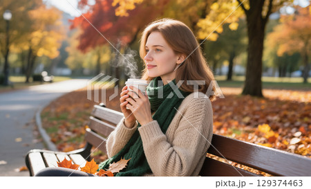 Young woman enjoying hot drink on bench in autumn park surrounded by colorful trees and fallen leaves 129374463