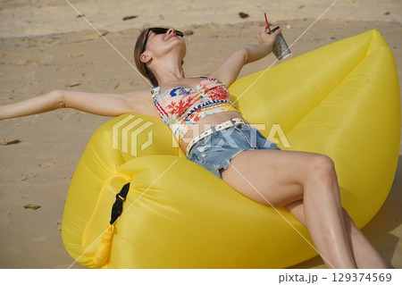 A young woman on the beach lounging on a yellow bean bag  129374569