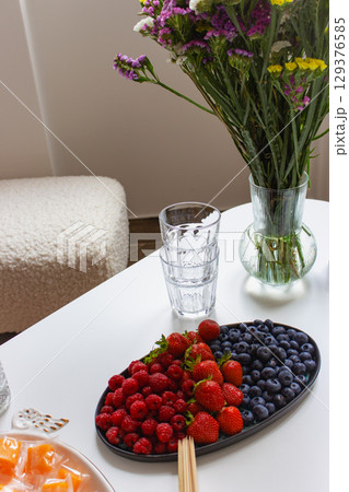 Blueberries, strawberries and raspberries on plate with wooden sticks on white table. Summer berries and flowers in vase. Summer harvest. Sweet berries and fruits. Healthy food. Natural lifestyle. Blueberries, strawberries and raspberries on plate with wooden sticks on white table. Summer berries and flowers in vase. Summer harvest. Sweet berries and fruits. Healthy food. Natural lifestyle. 129376585