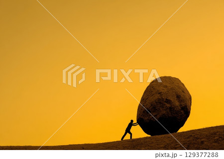 Silhouette of a Person Pushing a Boulder on a Mountain Slope at Dawn Against a Golden Sky 129377288