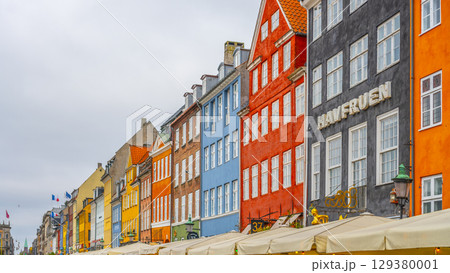 Vibrant, historic buildings line the canals of Nyhavn in Copenhagen, showcasing a mix of colors and architectural styles. Boats rest nearby, creating a lively atmosphere for visitors. 129380001