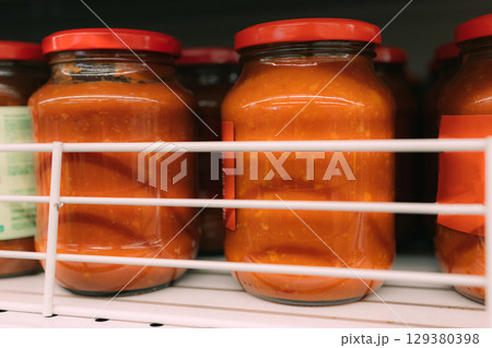 Glass jars with bright tomato sauce with red lids stand on the shelf of the store, Opititny view. Tomato sauce in a jar. Unevermag. 129380398