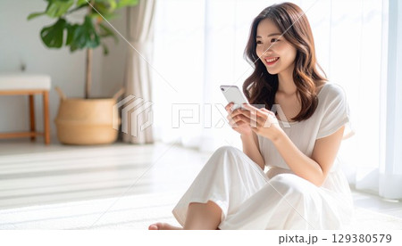 柔らかな日が差すリビングルームでスマートフォンを操作する若くて美しい日本人女性 129380579