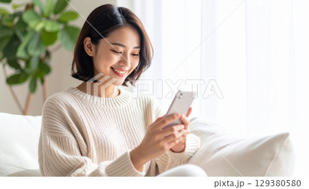 柔らかな日が差すリビングルームでスマートフォンを操作する若くて美しい日本人女性 129380580