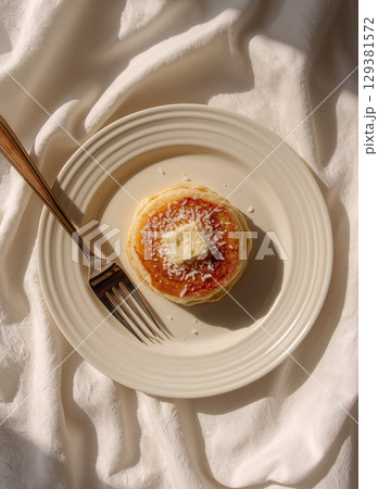 A classic pancake breakfast captured in a flat lay style. Pancakes with butter and syrup on a white plate with a gold fork, set against a softly wrinkled white cloth in bright morning light. A classic pancake breakfast captured in a flat lay style. Pancakes with butter and syrup on a white plate with a gold fork, set against a softly wrinkled white cloth in bright morning light. 129381572