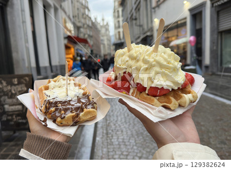 Enjoying two delicious belgian waffles topped with whipped cream, fresh strawberries, and chocolate sauce while exploring the charming streets of Brussels. 129382624