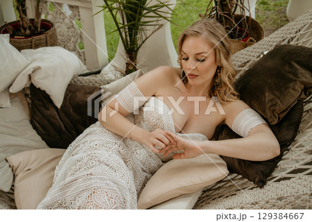 Beautiful slim blonde poses in boho wedding dresses and an embroidered corset on a summer terrace Beautiful slim blonde poses in boho wedding dresses and an embroidered corset on a summer terrace 129384667
