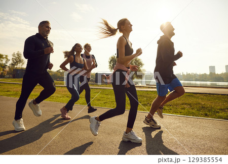 Group of sporty smiling people in sportswear jogging together in the park at sunset. Group of sporty smiling people in sportswear jogging together in the park at sunset. 129385554