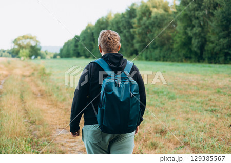 Rear view of blonde man with backpack standing in the countryside. Traveler. Summer time. Portrait of happy young man in casual clothing standing outdoor 129385567